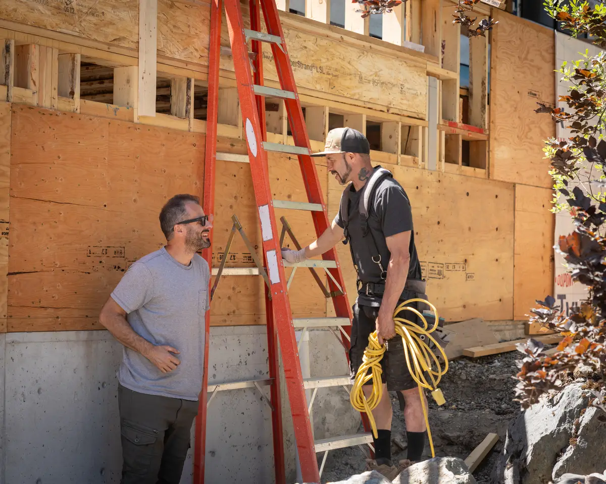 Two men beside a red ladder, one holding a coiled yellow cord, plywood wall behind
