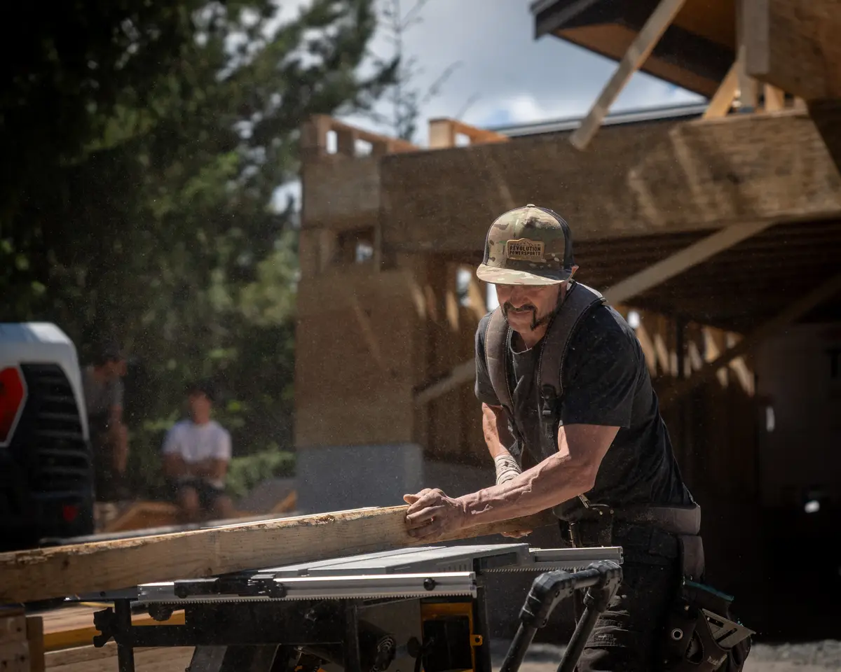A carpenter in a cap pushing a board through a saw, dust in the air, framing behind
