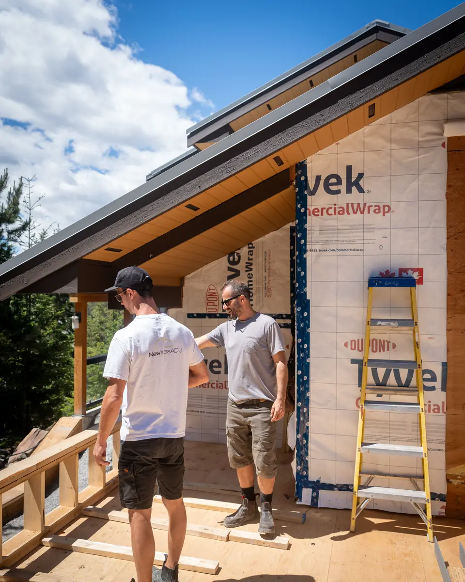 Two men on a deck under a wood soffit, house wrap on the wall, Newlines ADU on a white shirt
