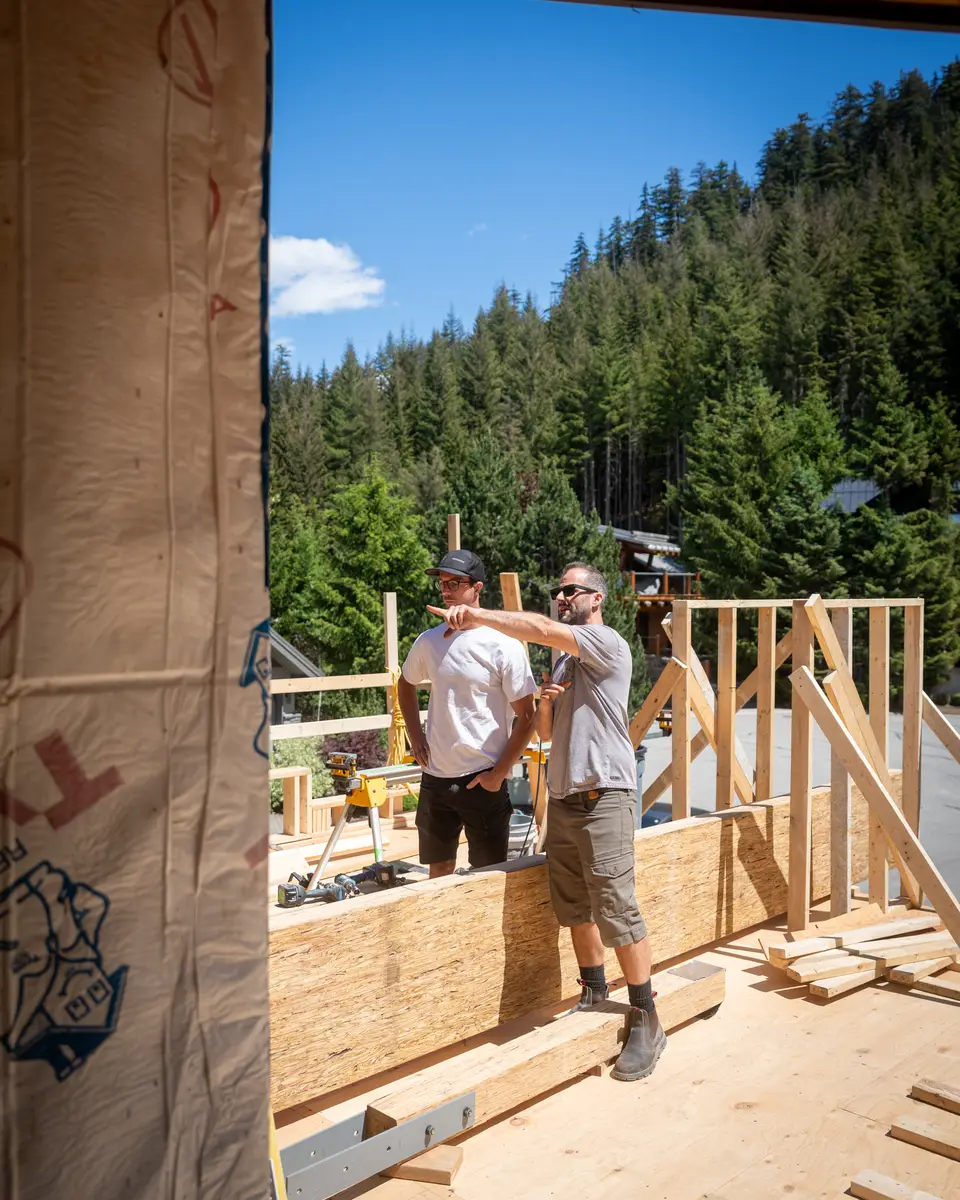 Two men on a plywood deck, one pointing past the studs, firs behind