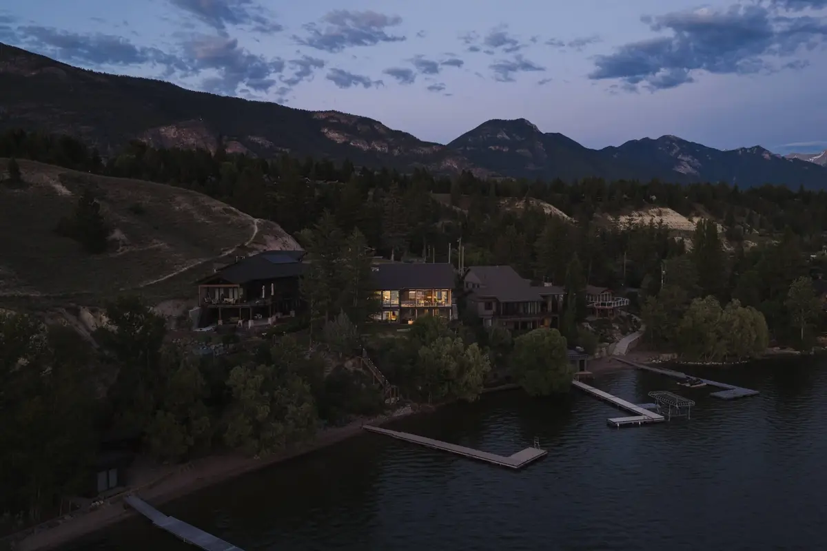 Dusk from the air over the lake, the lit house among trees above docks and shoreline