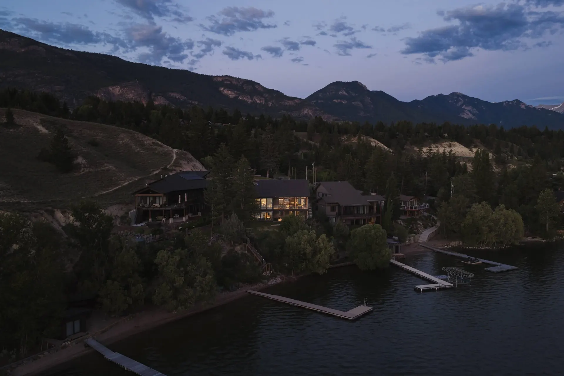 Dusk from the air over the lake, the lit house among trees above docks and shoreline