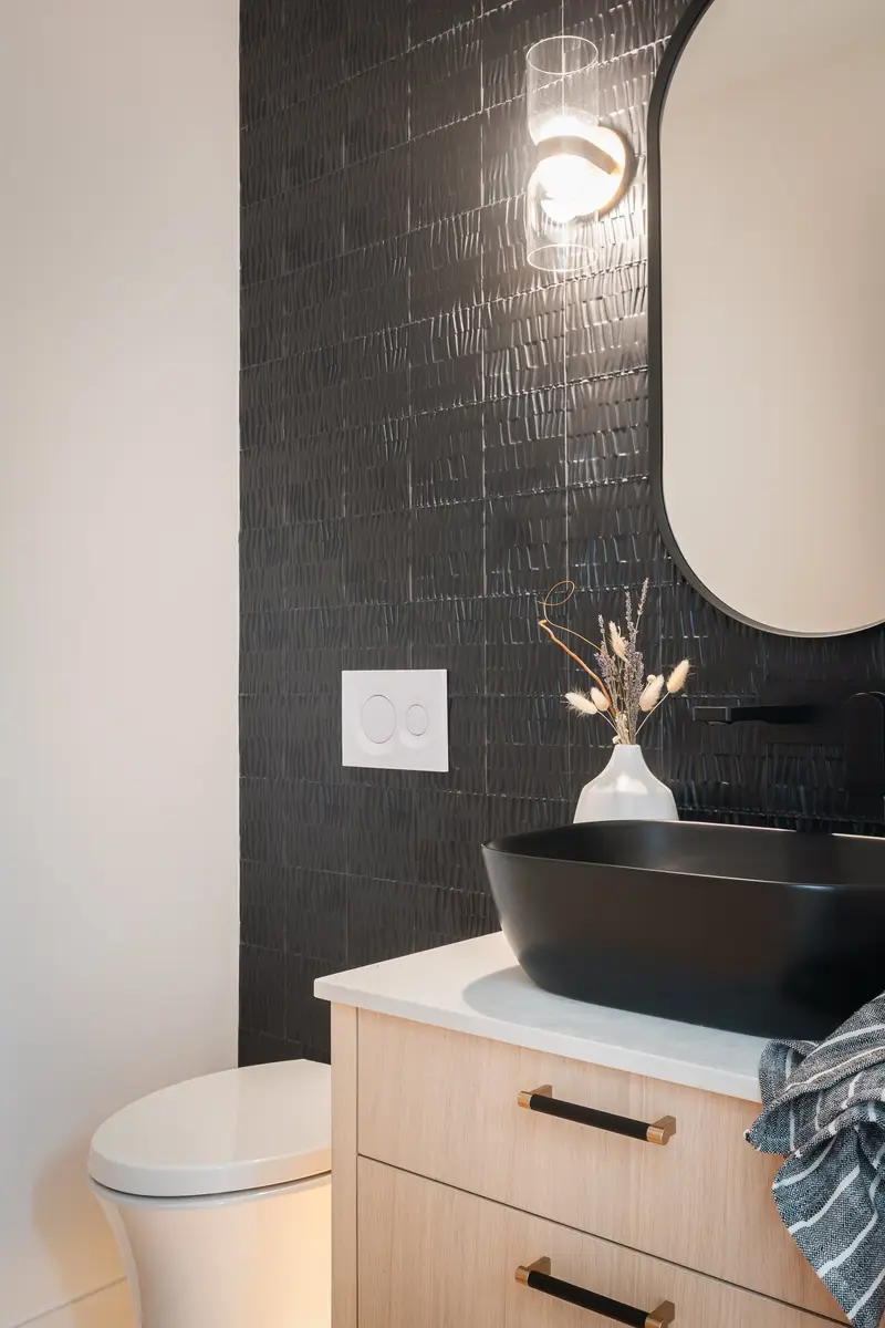 Powder room with a black vessel basin on a pale vanity, dark tile behind