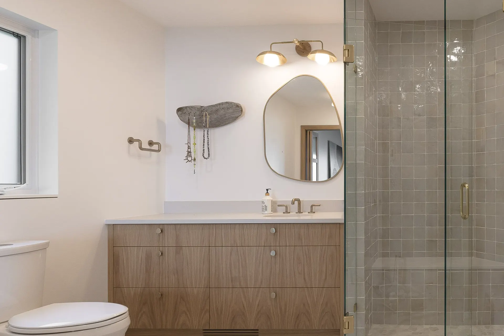 Vanity in wood grain with an irregular mirror and brass sconce, tiled shower behind glass