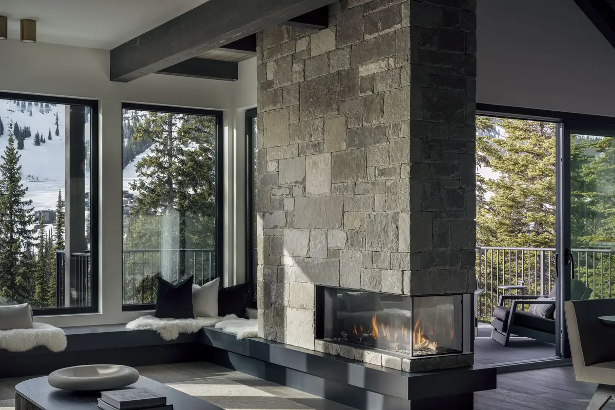 Stone chimney with a lit three sided fireplace, windows to snow on both sides