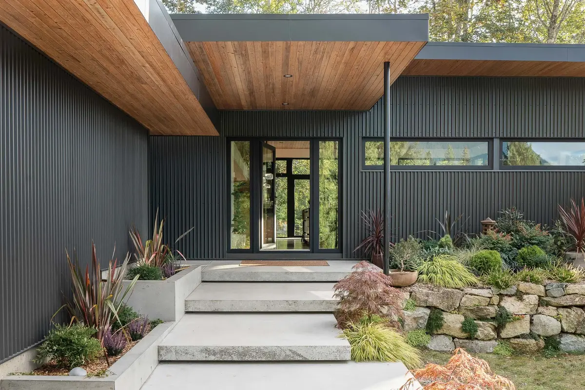 Entry under a deep wood soffit, dark corrugated metal cladding and concrete steps rising through planting