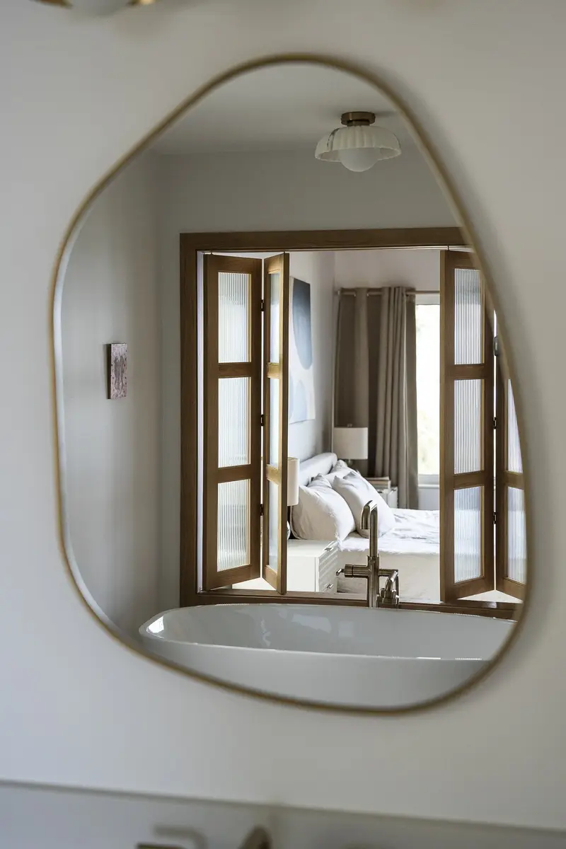 An irregular mirror reflecting the tub and reeded glass doors folded open onto the bedroom