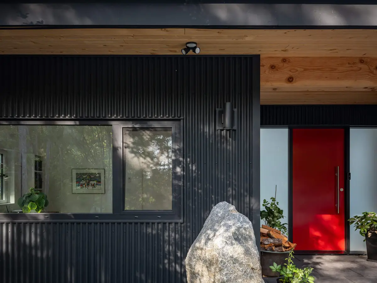 A red front door set into dark corrugated cladding beside a large boulder