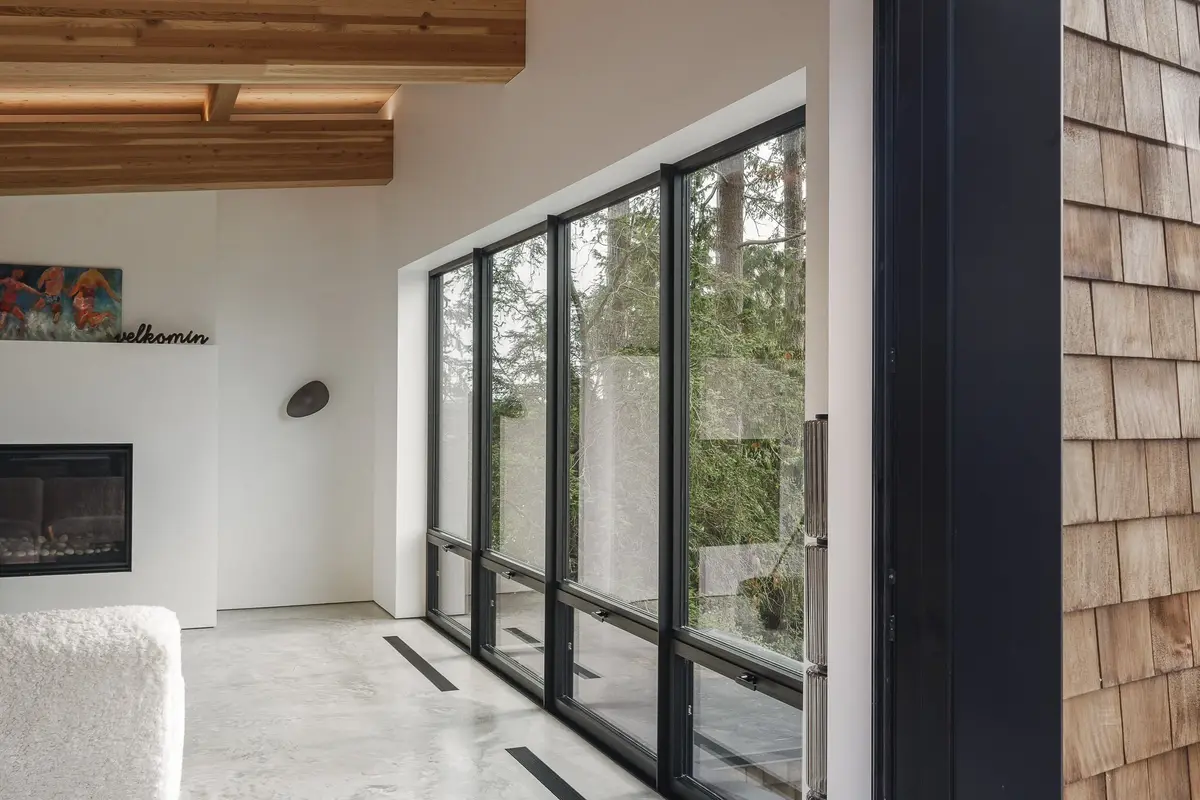 Window wall onto the forest beside a fireplace, concrete floor and beams overhead