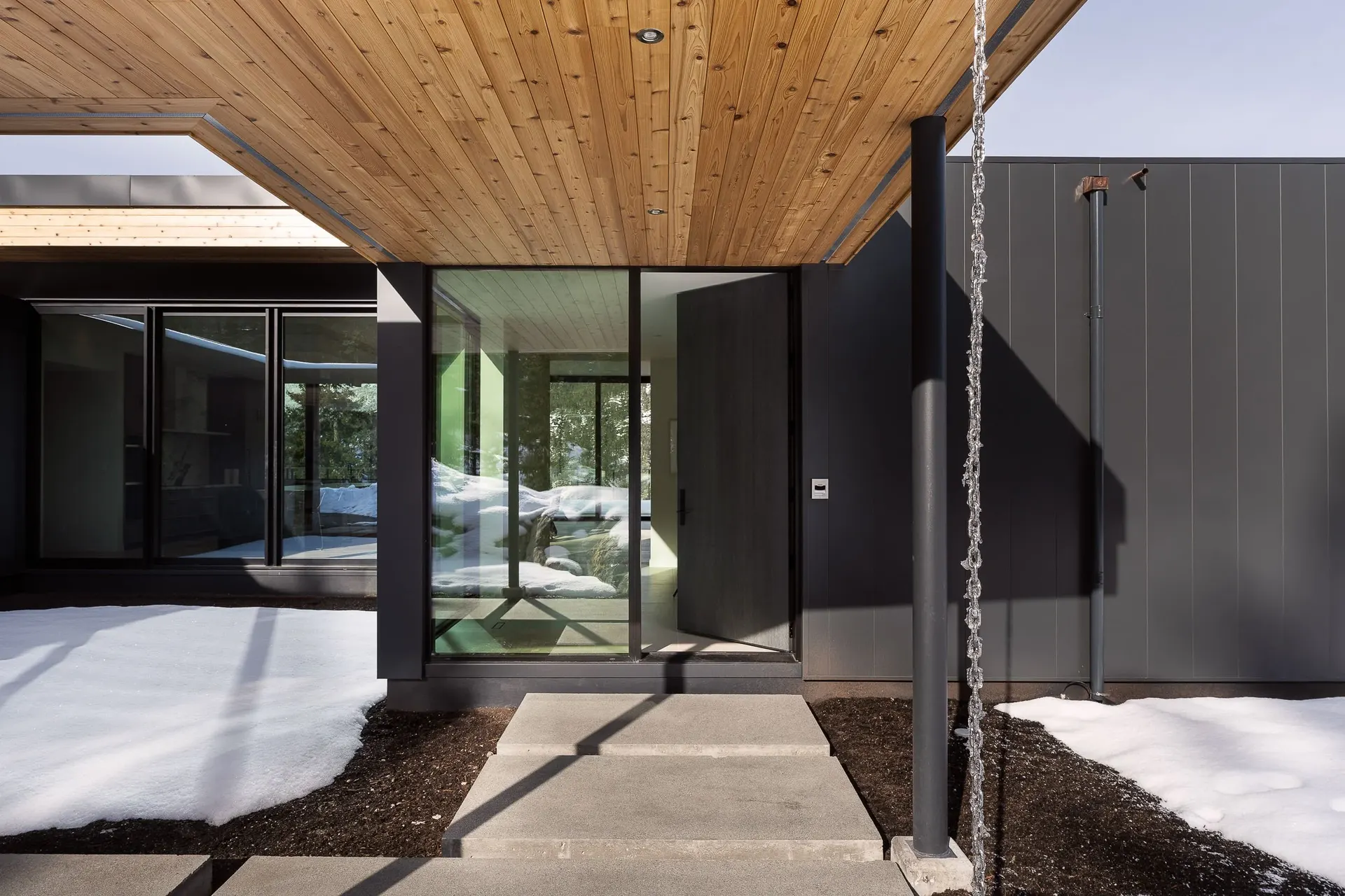 Covered entry walk on slim steel columns under a pale wood soffit, glazed entry beyond