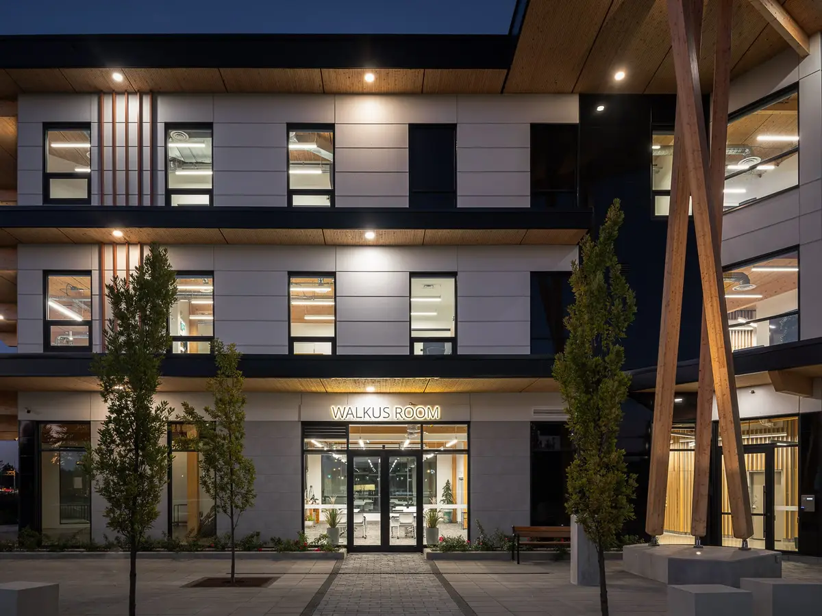 The entry elevation at dusk, lit floors behind glass and young trees on the plaza
