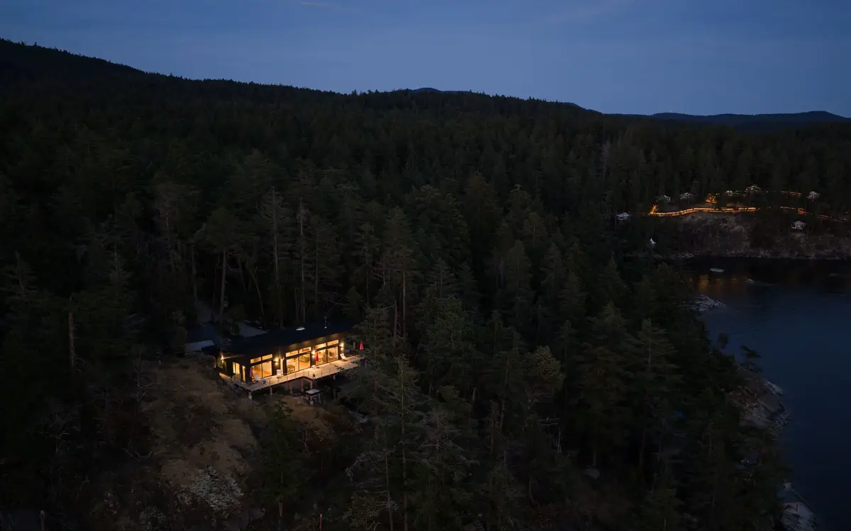 Twilight from above, the lit house small against the forested shore and dark water