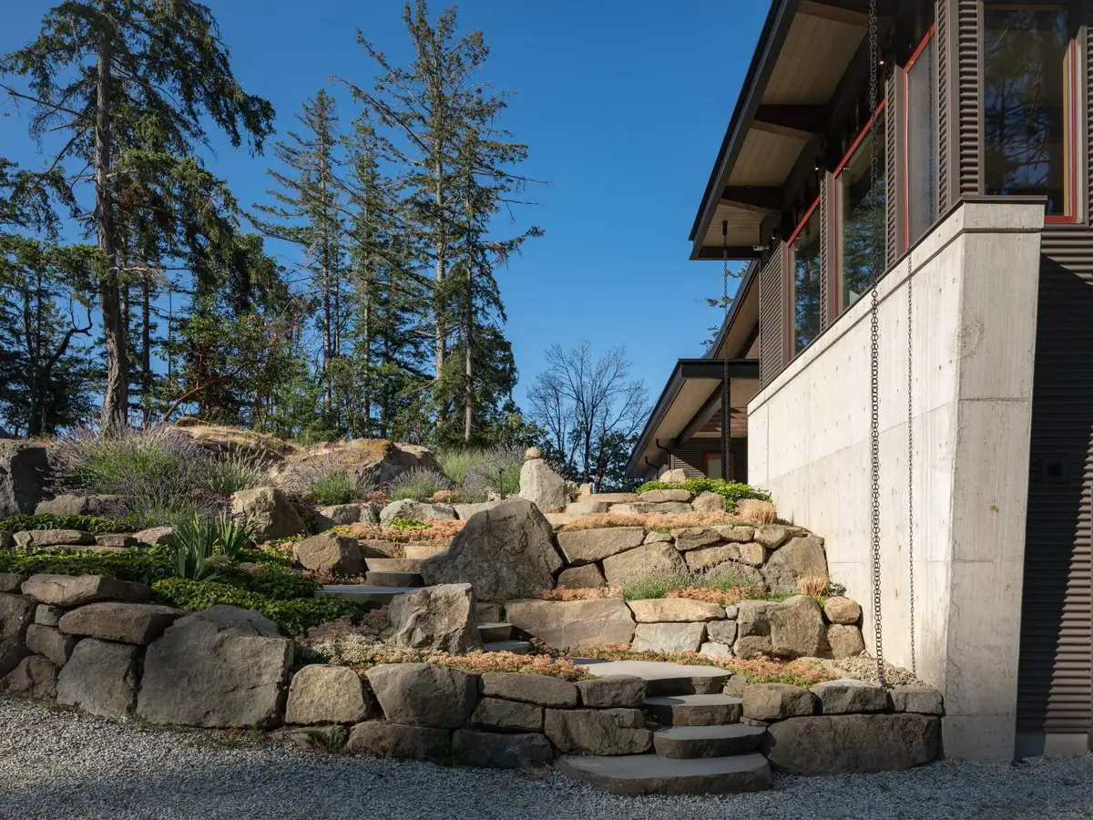 Stone steps rising through a rock garden beside a pale concrete wall and rain chains