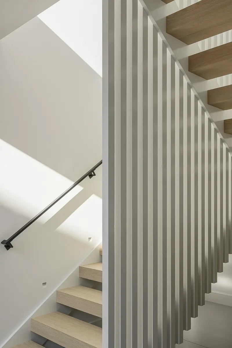 Open riser stair rising behind a white vertical slat screen, black handrail on the wall