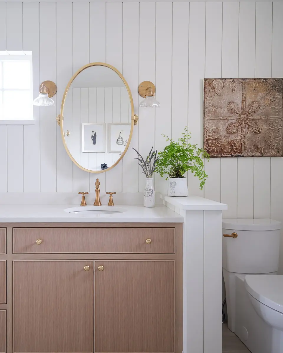 Vanity in pale wood with gold toned taps below an oval mirror, a framed print alongside