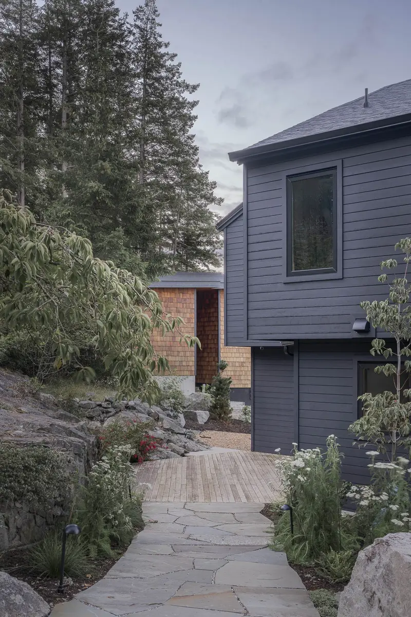 A dark clad house above a stone path, a shingled wing beyond and conifers behind