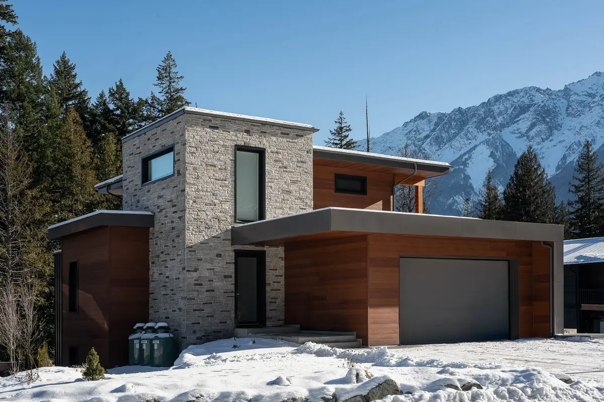 The house in snow below a mountain ridge, a stone volume beside horizontal wood cladding