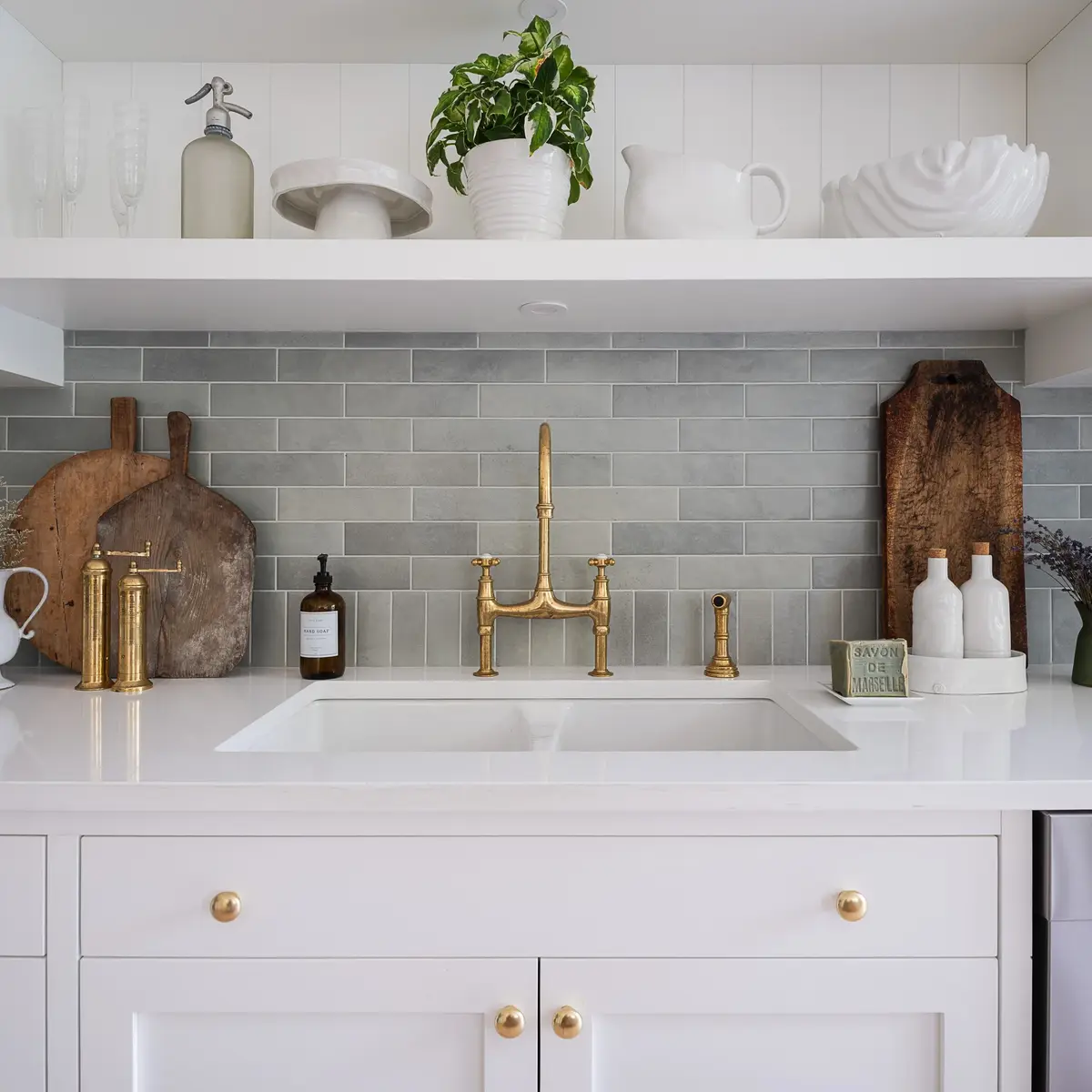 Undermount double sink and a tall gold toned tap against green tile, wood boards leaning either side