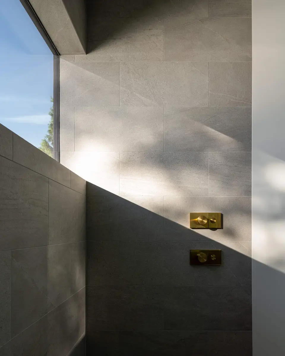 Grey tiled shower corner with brass coloured controls, daylight raking down the wall