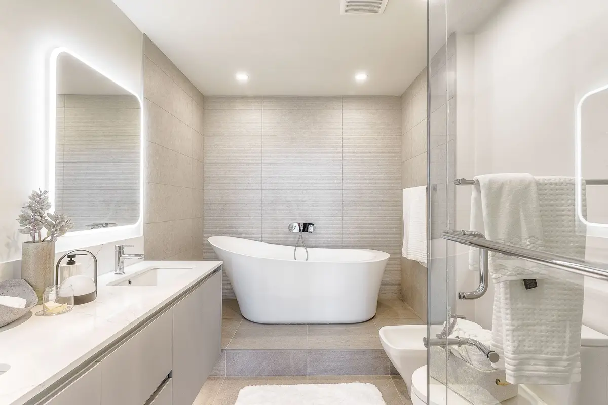 Freestanding tub against a tiled wall, a long vanity and lit mirror down one side