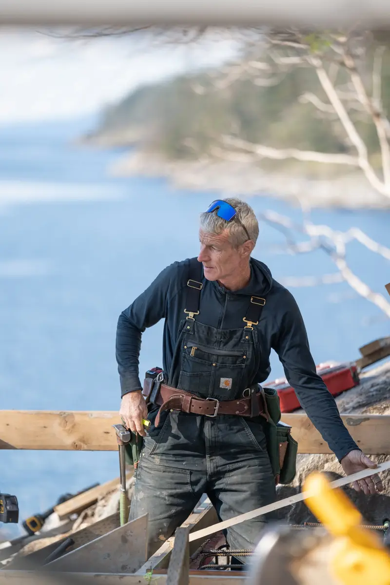 A builder in overalls with a tool belt, the water behind him