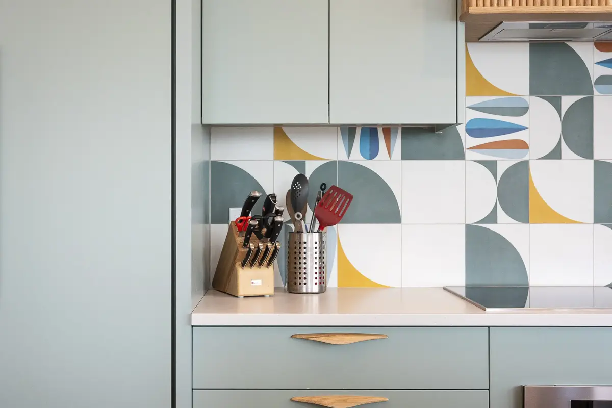 Geometric patterned tile behind a counter, knives and utensils above green drawers with wood pulls