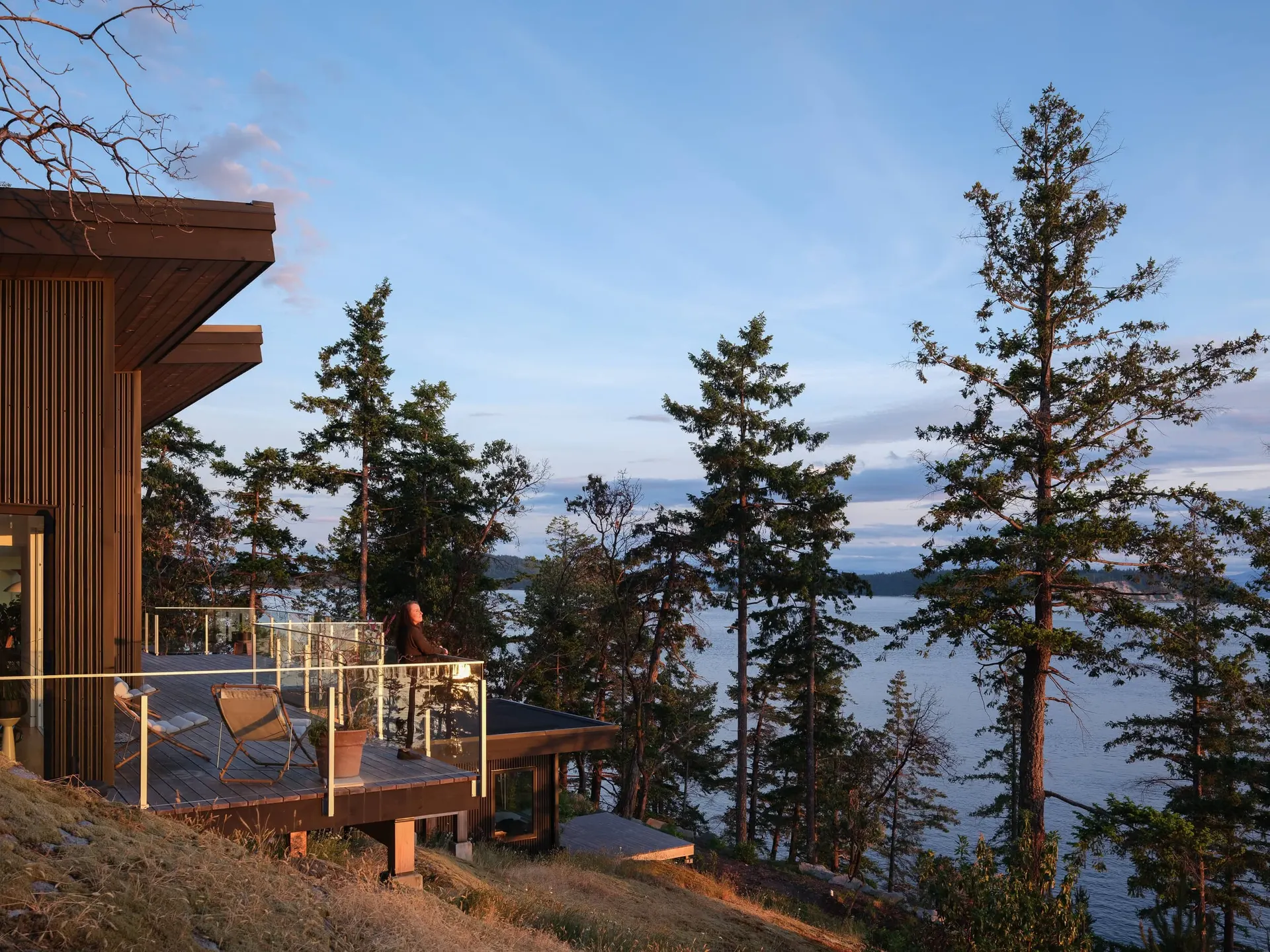 The house and its deck in evening light above the water, firs down the slope