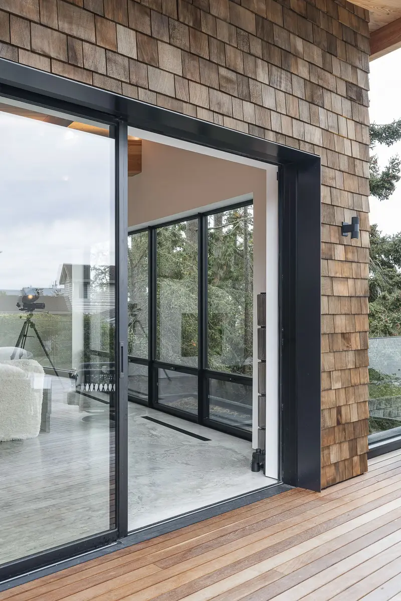 An open sliding door in a shingled wall, deck boards and glass railing outside