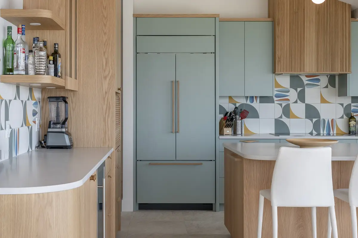 Tall wood cabinets and a green fronted fridge beside patterned tile, bottles on open shelves