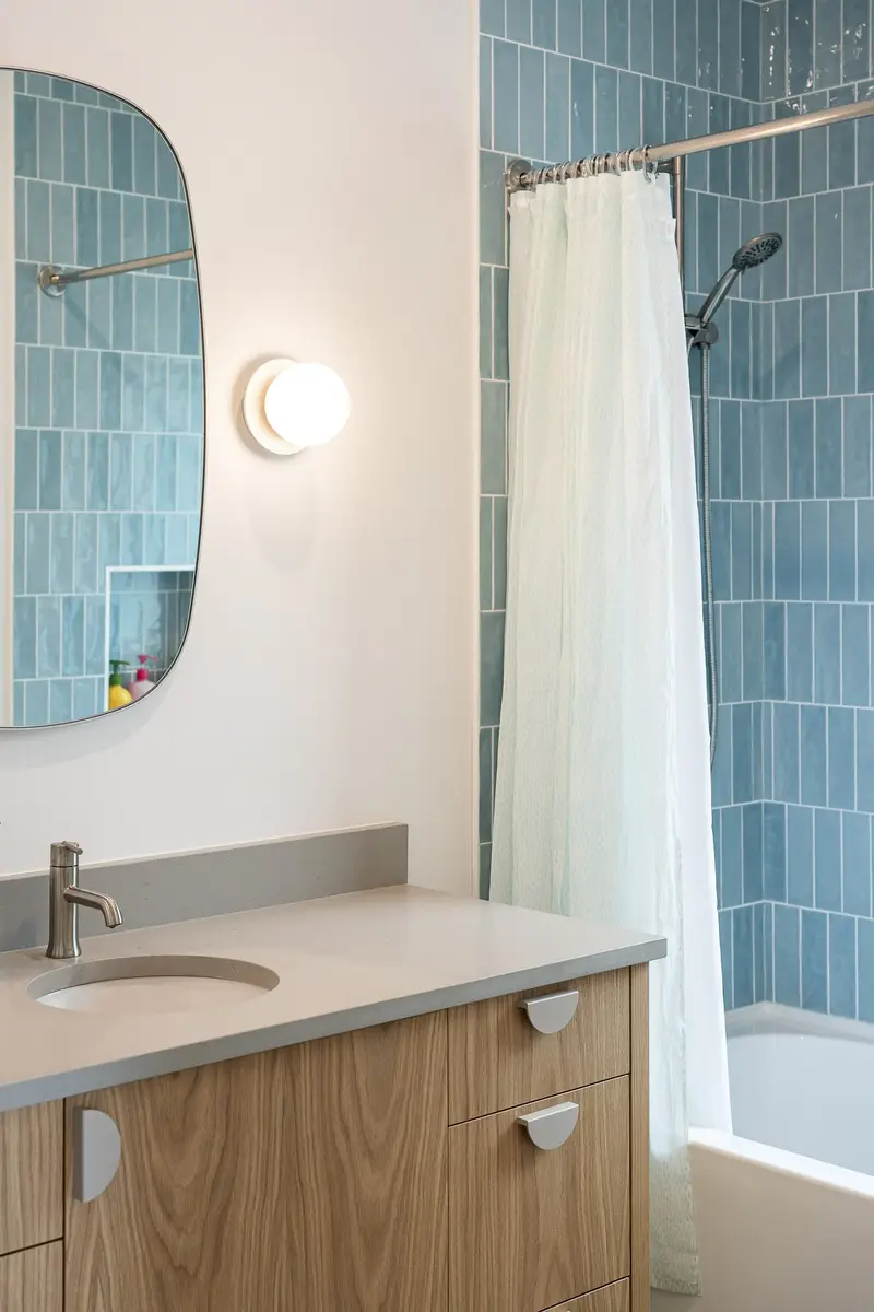 Blue tiled tub surround beside a wood vanity, a round wall light beside an irregular mirror