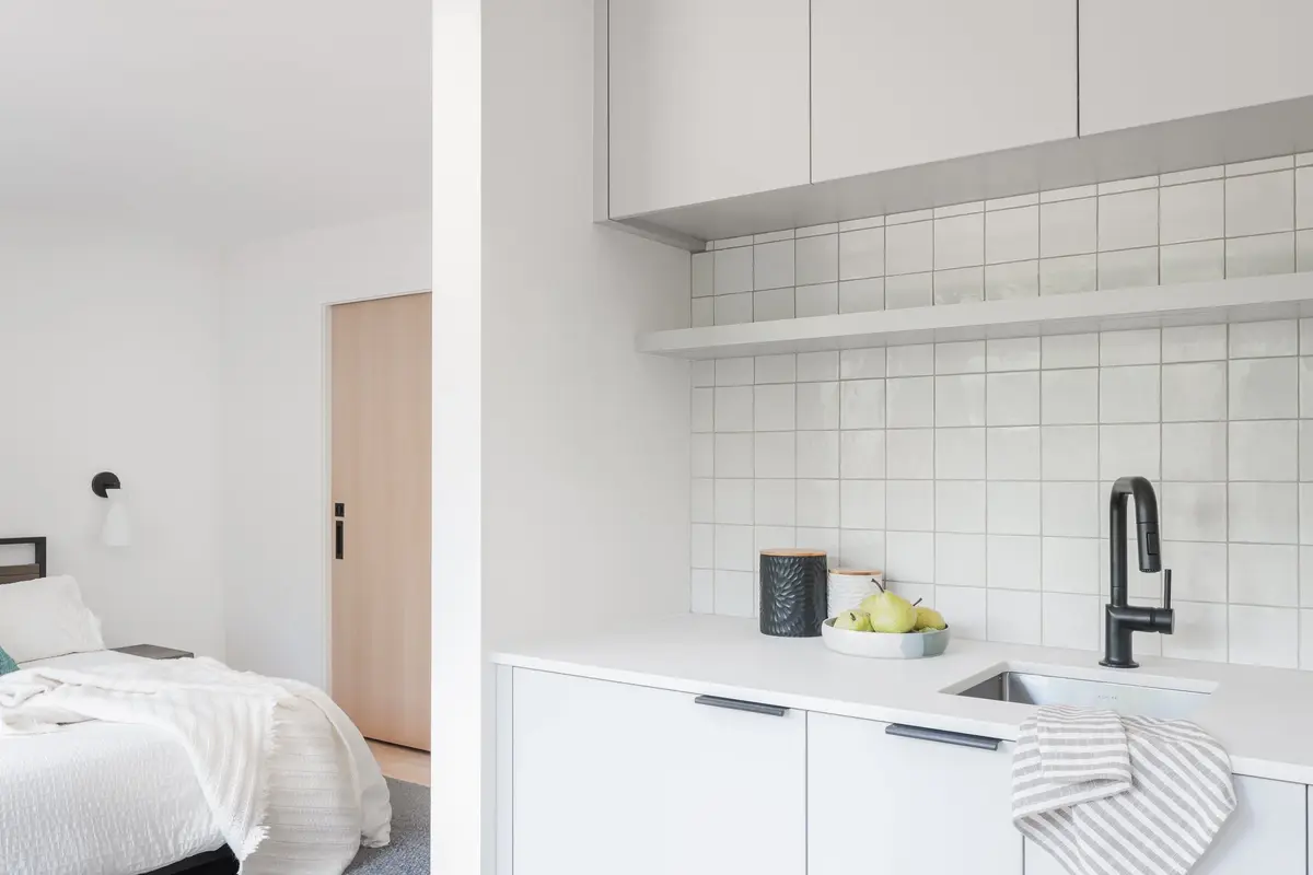 Bedroom through a doorway at left, a white tiled kitchenette counter at right