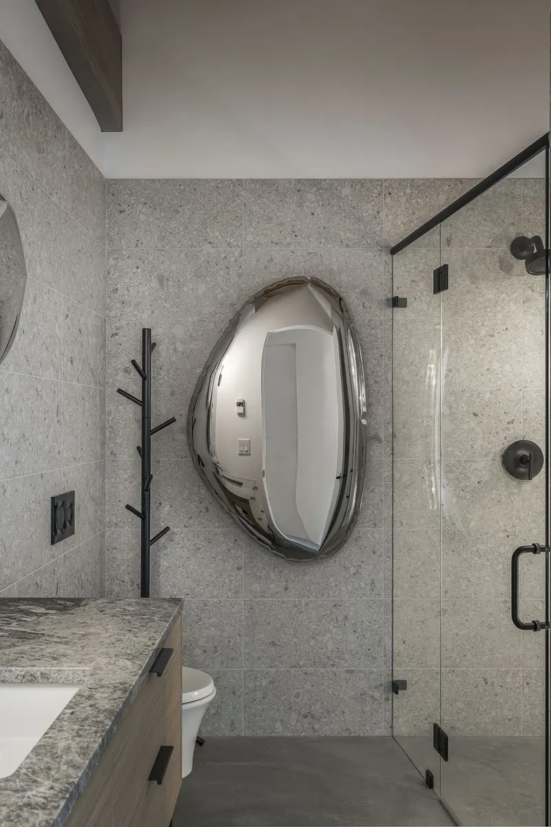 An irregular mirror on a speckled tile wall beside a glass shower enclosure
