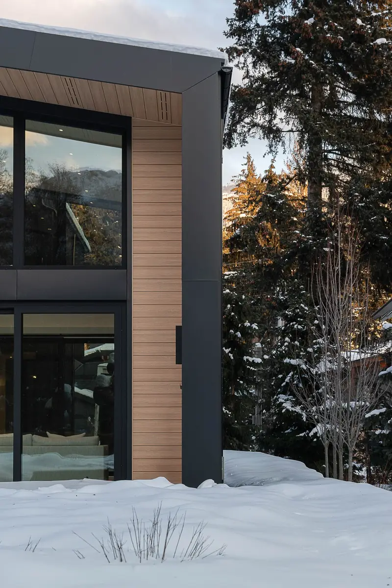 A wood clad corner meeting dark cladding, glazing reflecting the trees above deep snow