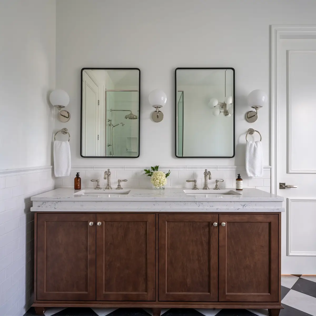 Double vanity in dark wood below two black framed mirrors and globe sconces