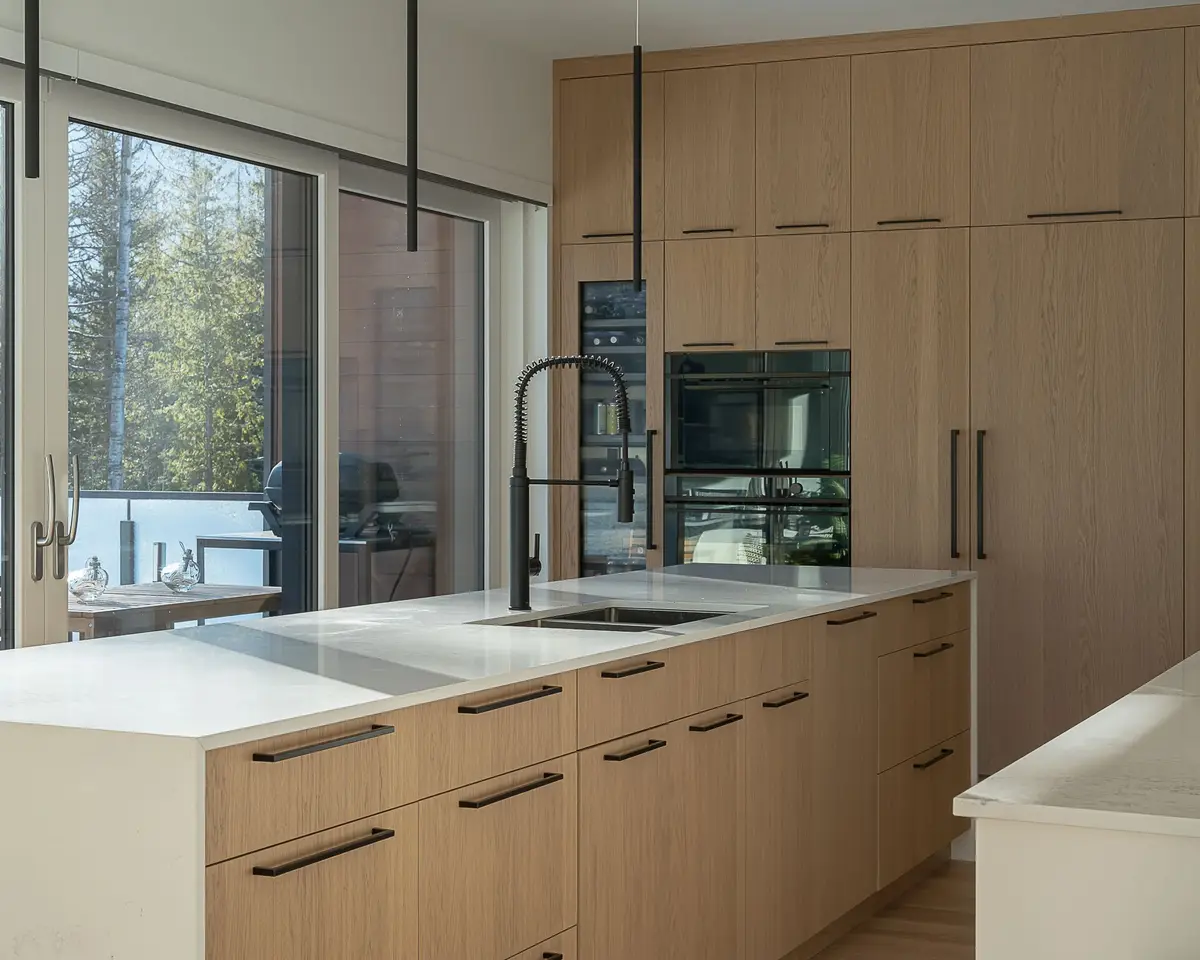 Island sink and black tap below wood cabinetry with built in ovens, trees outside