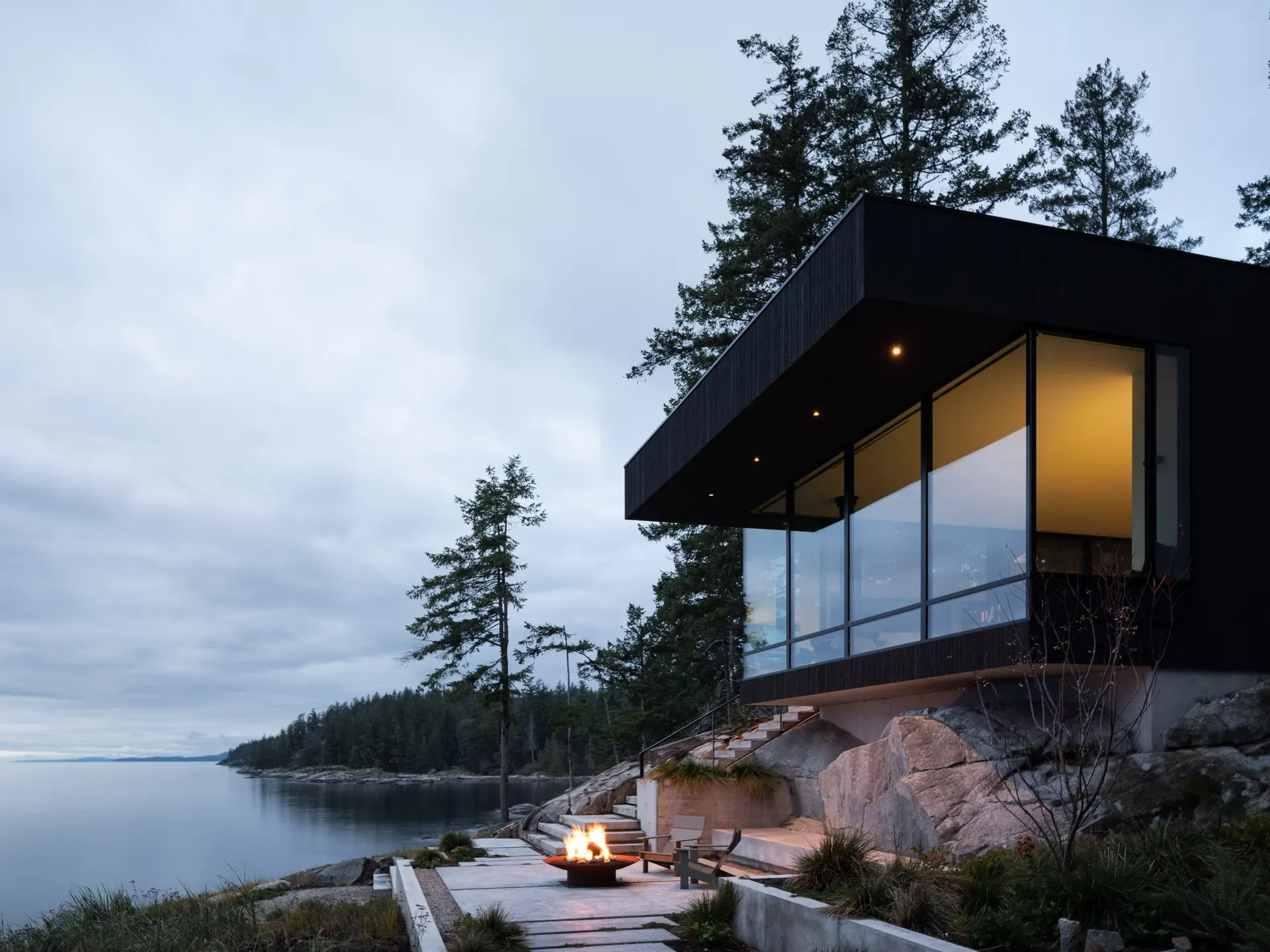The house at dusk over the shoreline, a fire burning on the terrace beside the water