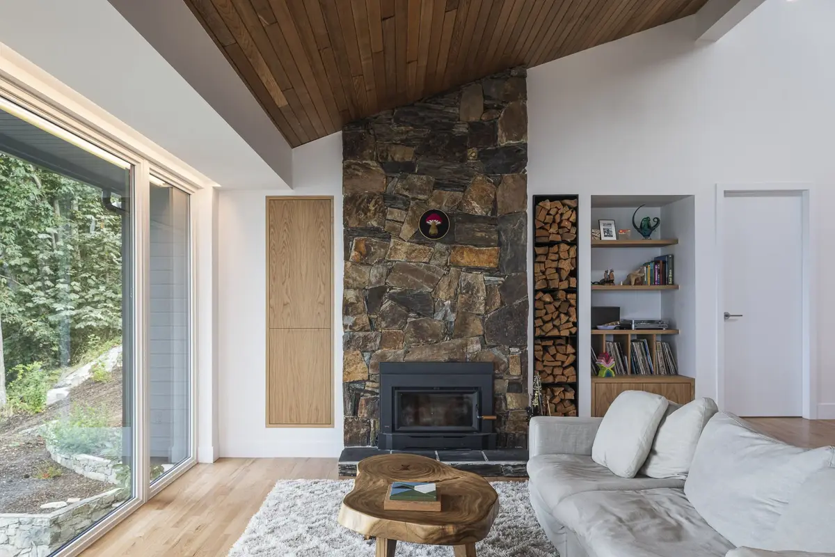 Living room under a sloped wood ceiling, stone chimney and sliding glass onto the forest