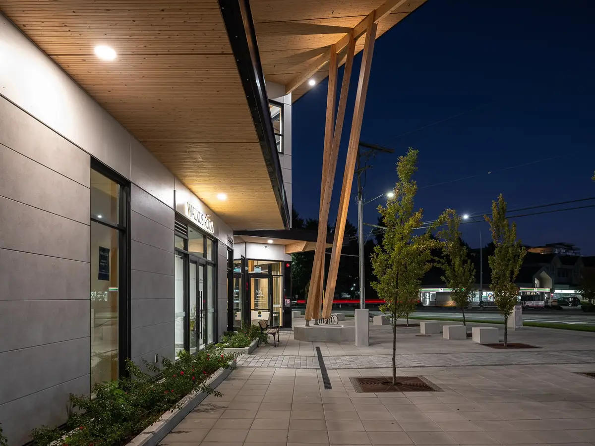 Along the covered walk at dusk, raking timber columns and the lit soffit above the paving