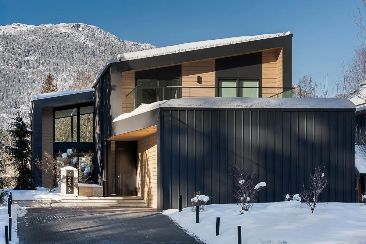 Street elevation above a cleared drive, dark cladding below and wood siding above, mountains beyond
