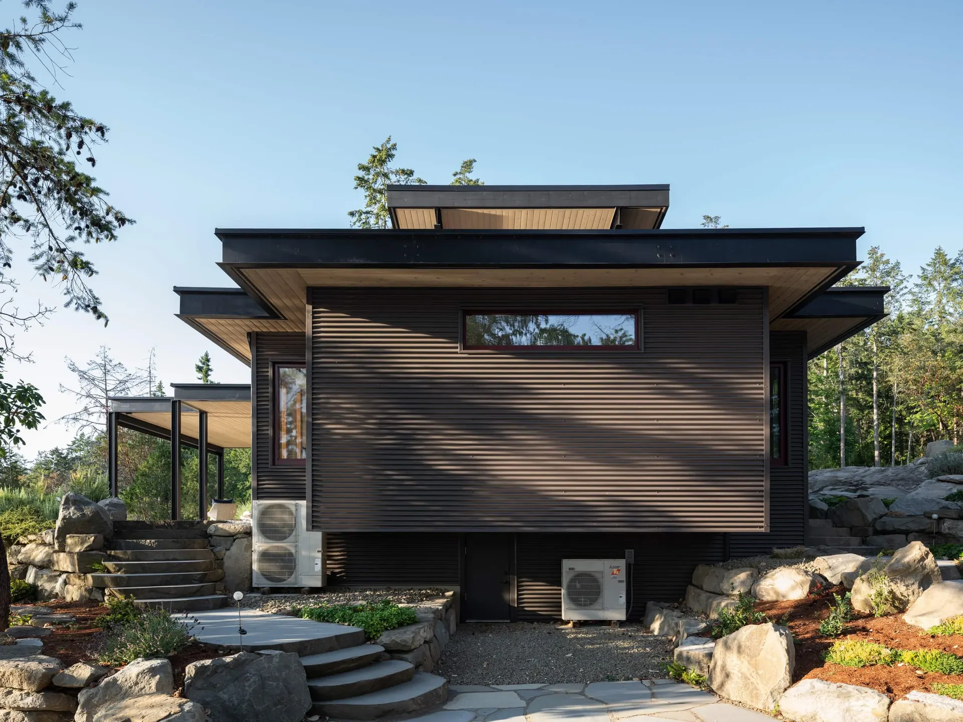 Stacked shed roofs over ribbed cladding, stone steps climbing between boulders