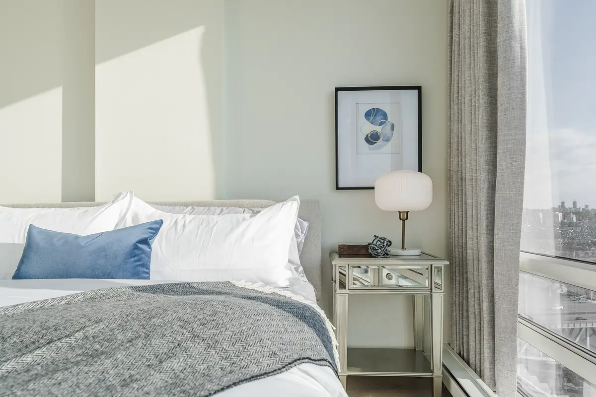 Bed made in white and blue beside a mirrored nightstand, a lamp and a framed print