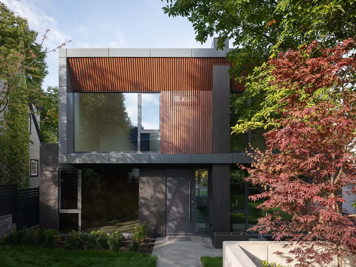 Two storey house behind a maple, dark panels below a wood slat screen and glazing