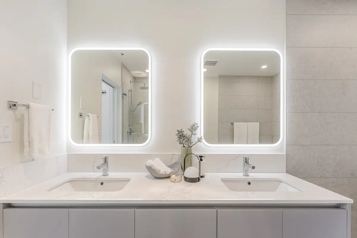 Two lit mirrors above a double vanity, a shower reflected in the left one