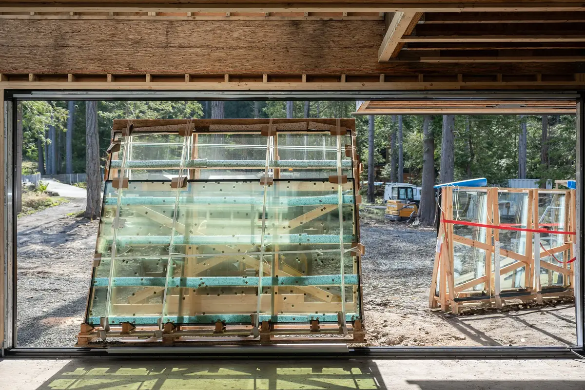 Crated glass units standing inside an unfinished opening, trees and a loader beyond