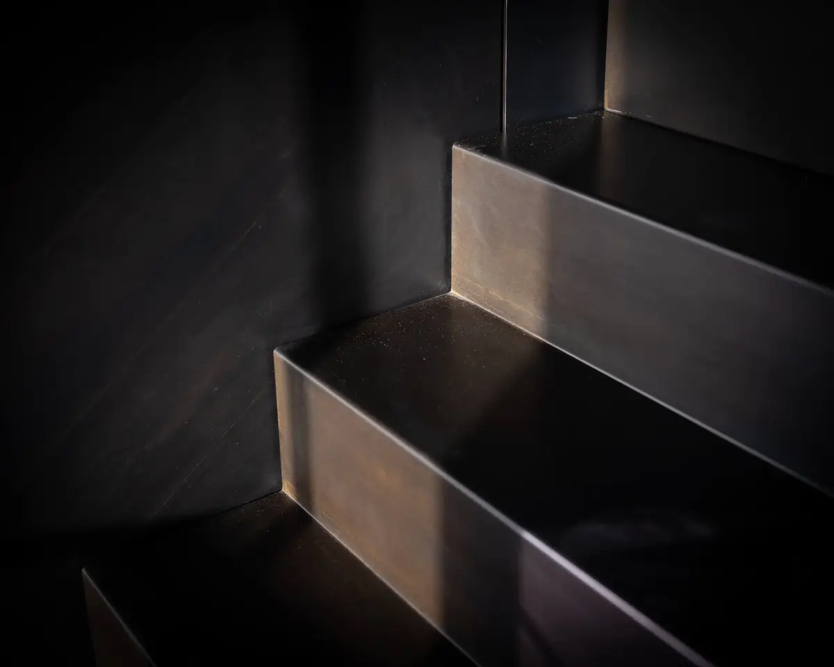 Folded dark metal treads meeting the stair guard, light along each nosing