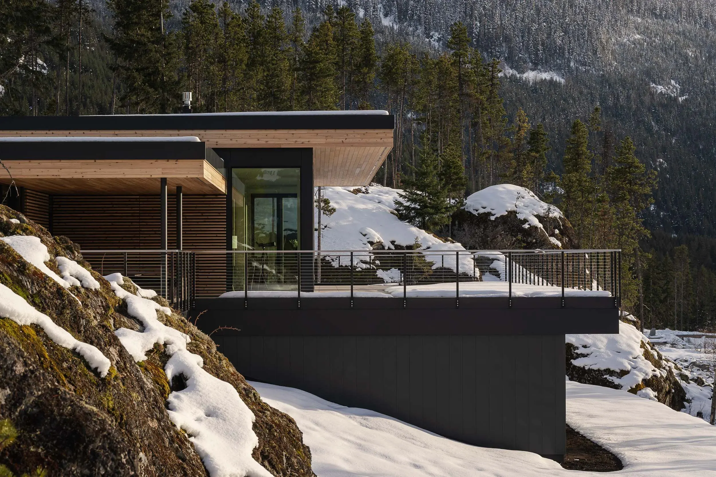 The house along the rock outcrop, dark cladding under a pale wood soffit with snow below