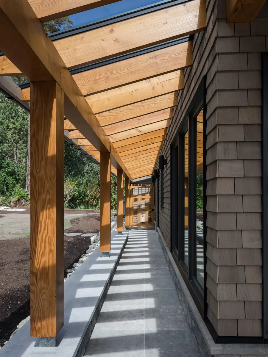 Timber posts carrying a glazed canopy along a shingled wall, shadows across the walk