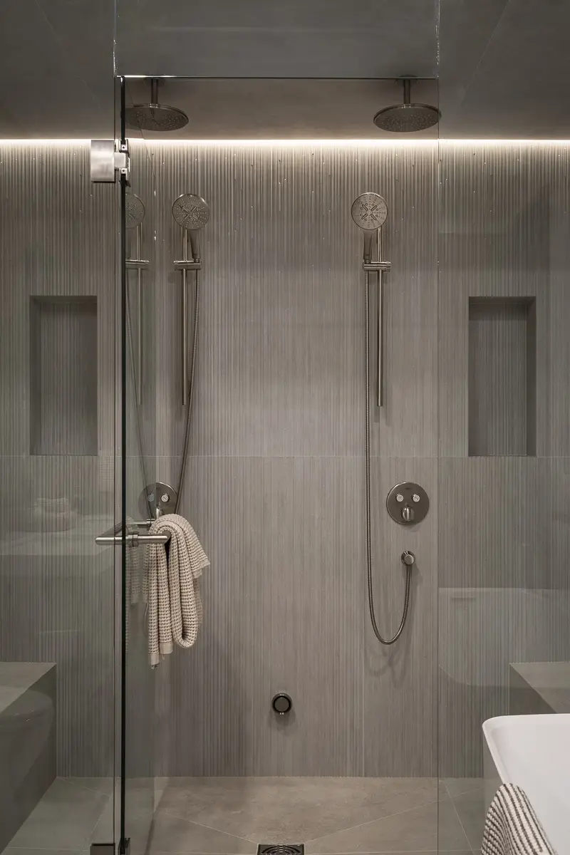 Two rainheads and handshowers in a tiled shower behind clear glass, a towel hanging