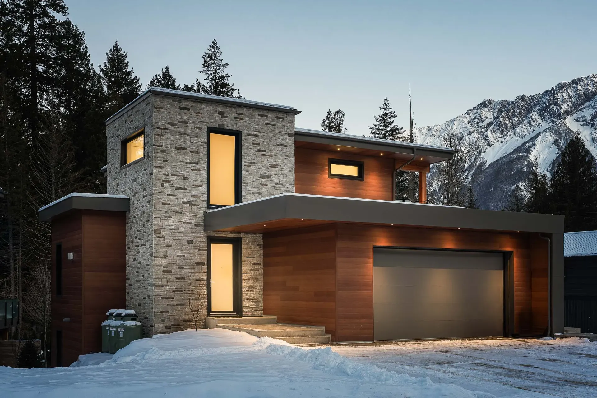 The house at dusk against the mountain, lit windows in the stone and wood elevations