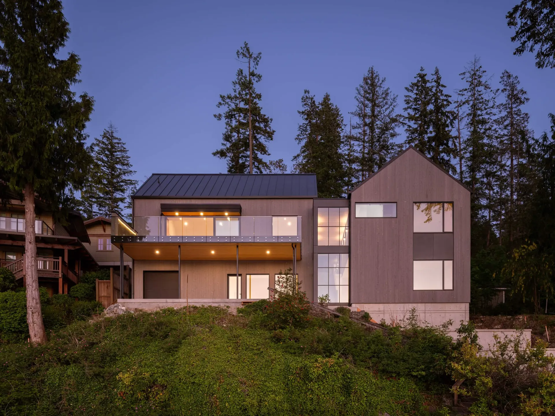 The house at twilight, deck and windows glowing between the trees above the slope